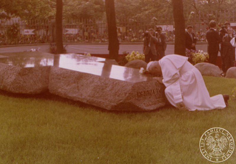 Jan Paweł II modli się przy grobie ks. J. Popiełuszki. Za nim grupa reporterów, fragment ogrodzenia kościoła pw. św. Stanisława Kostki. Za ogrodzeniem zebrany tłum. 13 VI 1987 r Sygnatura: IPNBU-7-14-11-12. Autor: Nieznany.