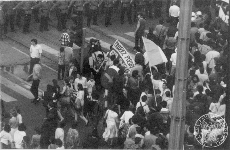 Szpaler milicjantów oddziela demonstracje pielgrzymów wracających ze spotkania z Janem Pawłem II na krakowskich błoniach. Widoczna flaga z napisem: Młodzież pozdrawia Ojca Świętego, 10 VI 1987 r., sygn. IPN Kr 056/105