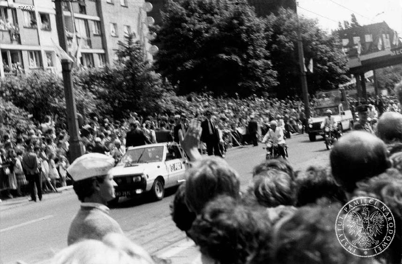 Jan Paweł II w papamobile, w asyście milicjantów na motocyklach i samochodu ochrony, przejeżdża ulicami Szczecina pozdrawiany przez wiernych. 11 VI 1987 r. IPN Sz 706/23 t. 1-2.