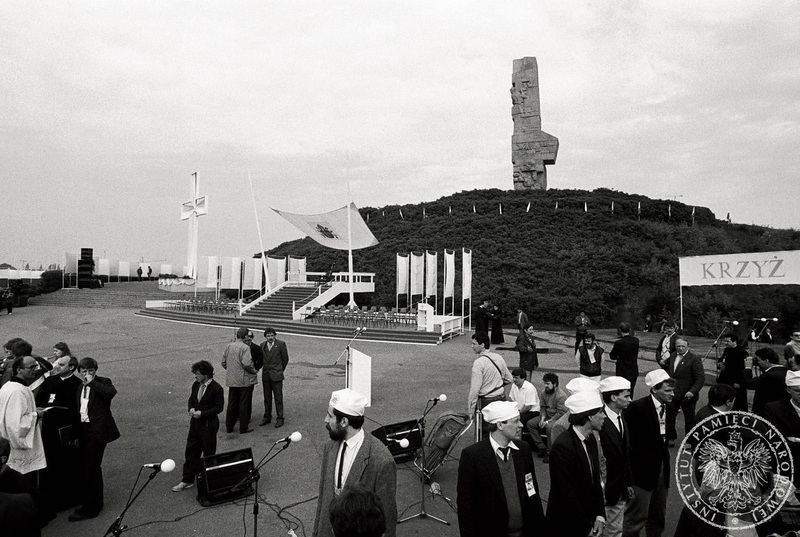 Podest i krzyż ustawione pod pomnikiem Bohaterów Westerplatte, po prawej członkowie Kościelnej Służby Porządkowej w oczekiwaniu na Ojca Świętego. 12 VI 1987 r. Sygnatura: IPNBU-7-7-8-180